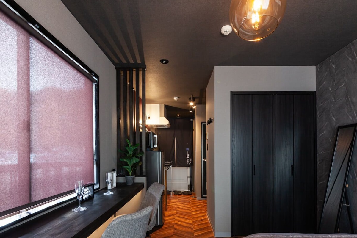 Una habitación de hotel moderna con suelo de madera en espiga, un ventanal grande, una cama, un escritorio con una silla y una lámpara colgante.