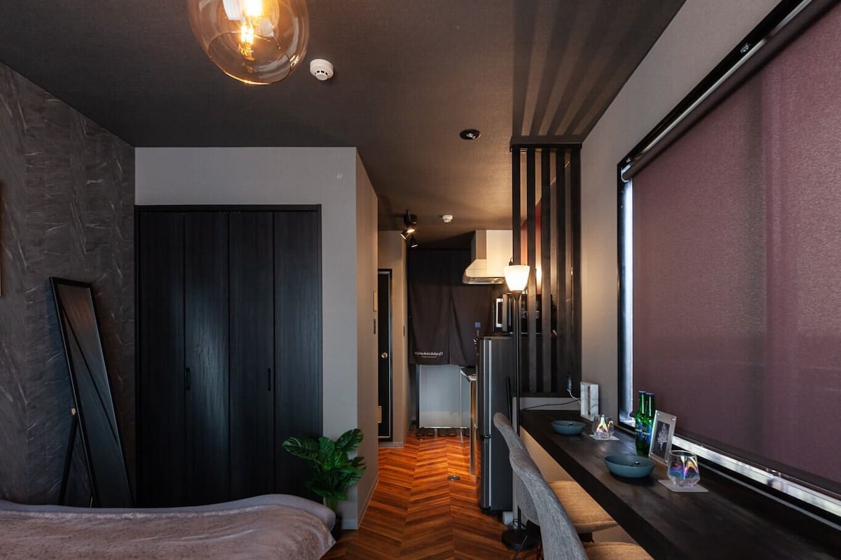 Una habitación de hotel moderna con piso de madera tipo espiga, una cama, un escritorio con silla, un ventanal grande y una lámpara fijada a la pared.