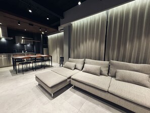 Uma sala de estar moderna com um sofá seccional, uma área de jantar com mesa e cadeiras de madeira e uma cozinha ao fundo.