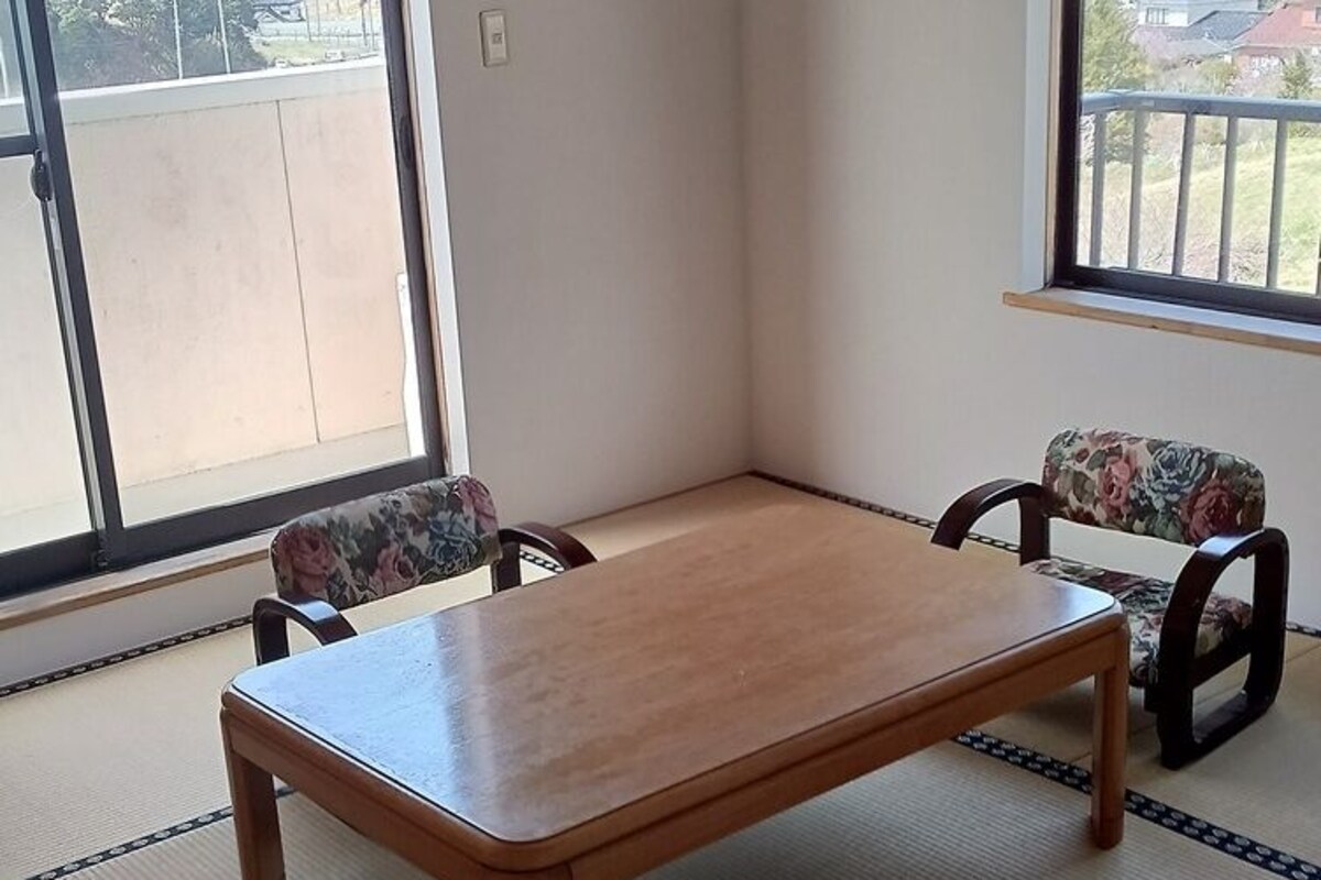 Une pièce avec un sol recouvert de tatamis, une table en bois, deux chaises et de grandes fenêtres donnant sur un paysage de montagnes et de maisons.