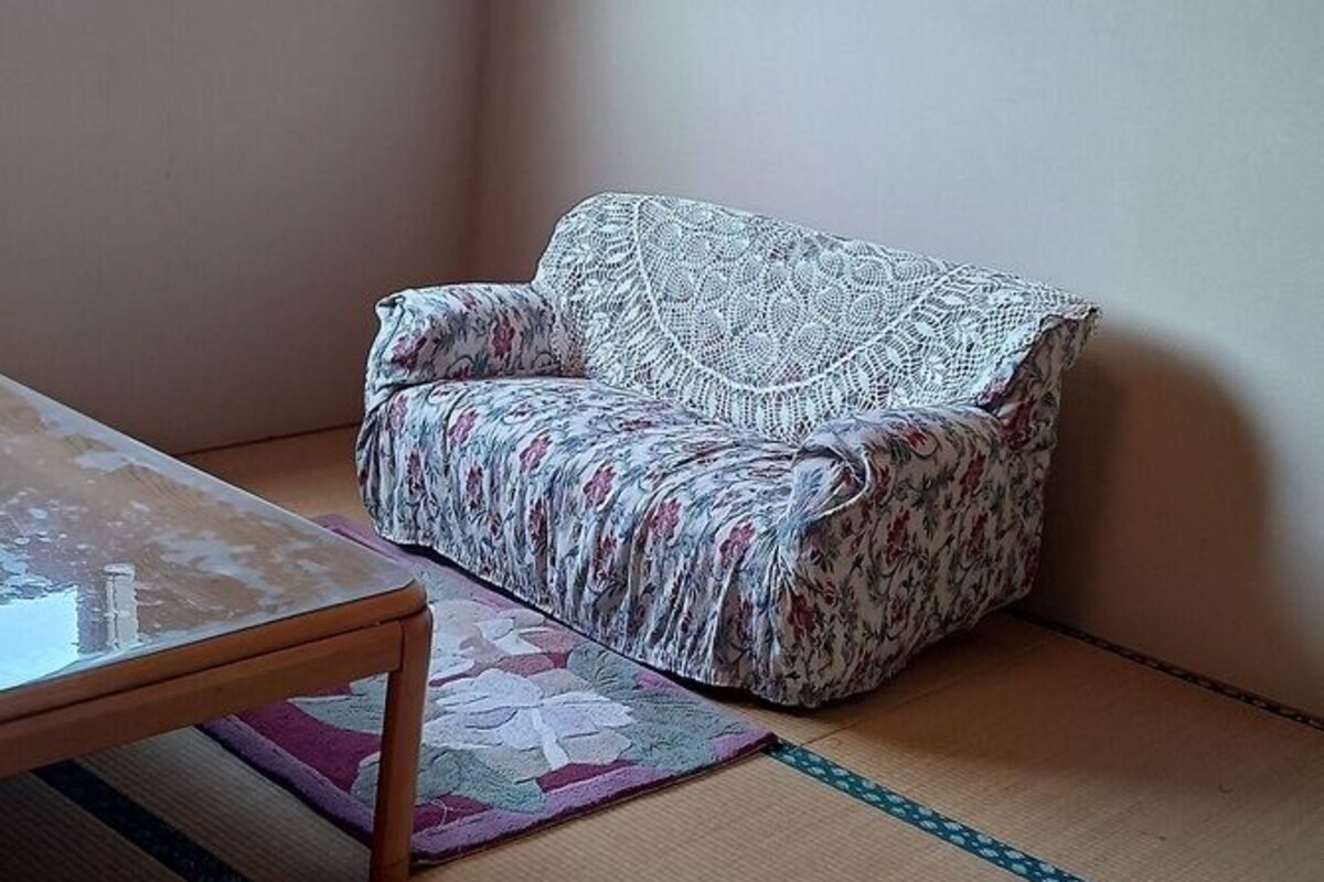 Une pièce avec un sol recouvert de tatamis, un canapé à motifs floraux, une table et un climatiseur fixé au mur.