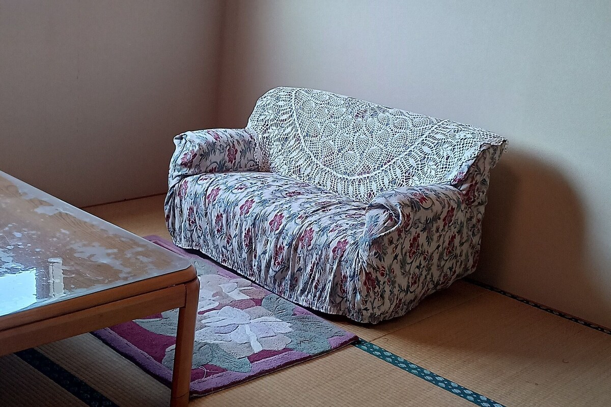 Une pièce avec un sol recouvert de tatamis, une petite table en bois, un canapé recouvert d’un tissu à motifs floraux, un climatiseur fixé au mur et une horloge ronde.