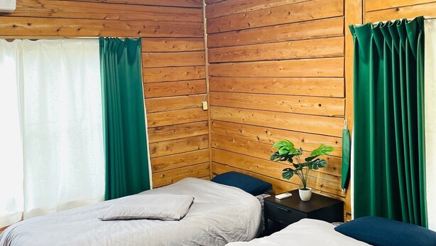 A room with two beds, wooden walls, a small table with a plant, and a ceiling light. - Wood Retreat (Miyazaki)