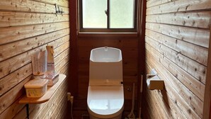 A small, wooden cabin-style bathroom with a toilet, a window, and a shelf with toiletries. - Wood Retreat (Miyazaki)