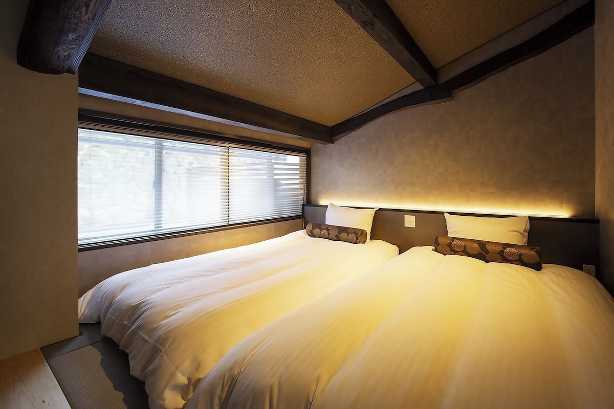 A bedroom with a large bed, a window with blinds, and a headboard with built-in lighting.