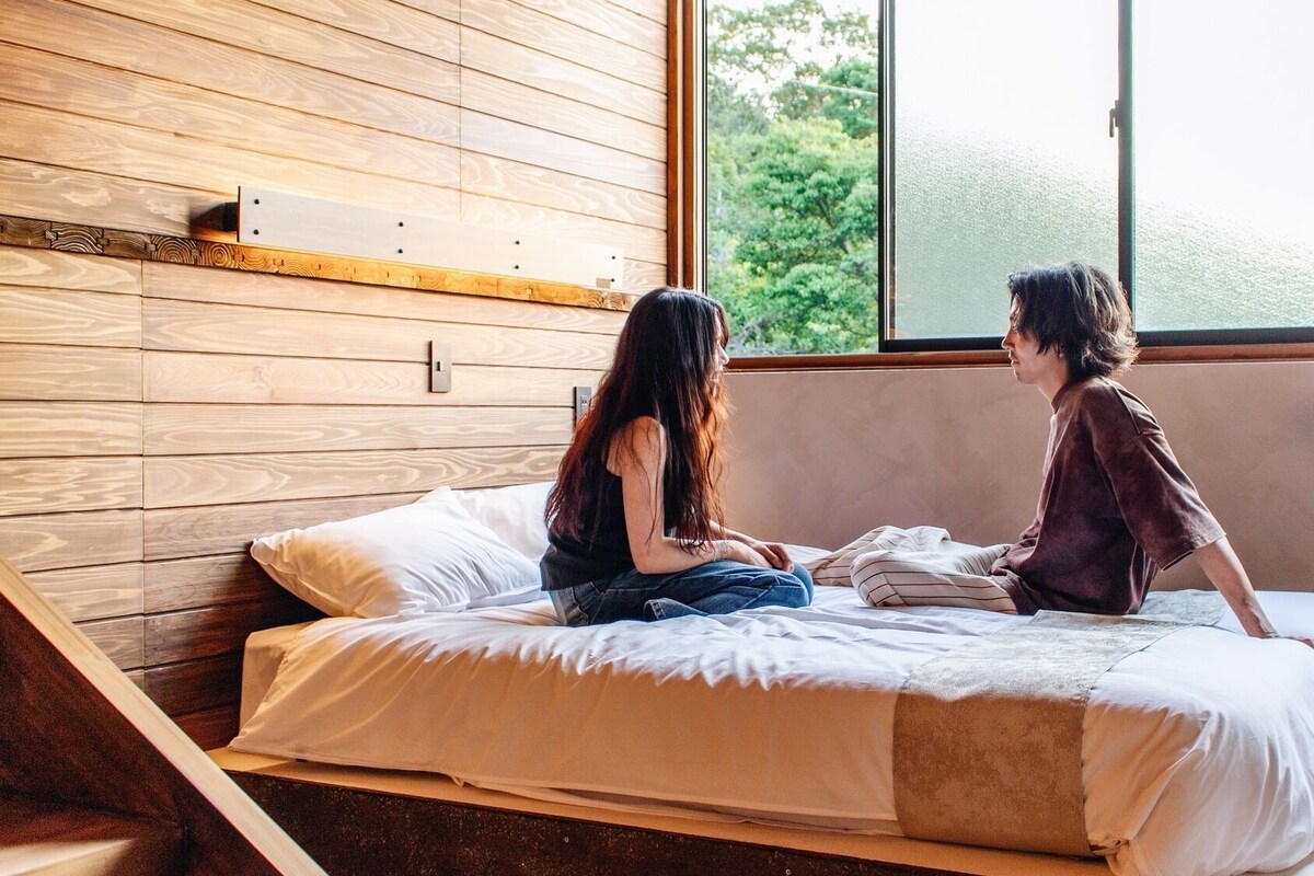 A room with a bed, wooden panel walls, a window with a view of trees, and a light switch on the wall.