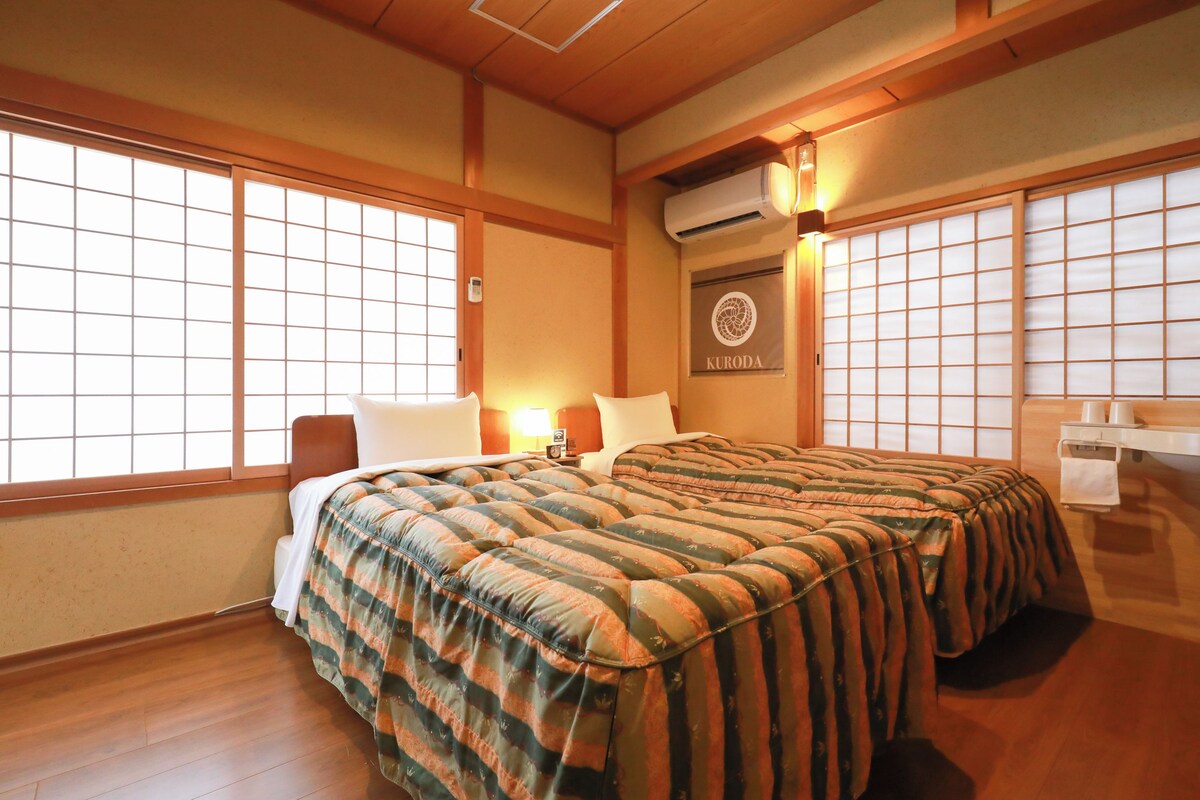 Una camera da letto in stile tradizionale giapponese, con pavimento in tatami, un letto grande con biancheria a righe, una porta scorrevole shoji e un condizionatore d'aria a parete.