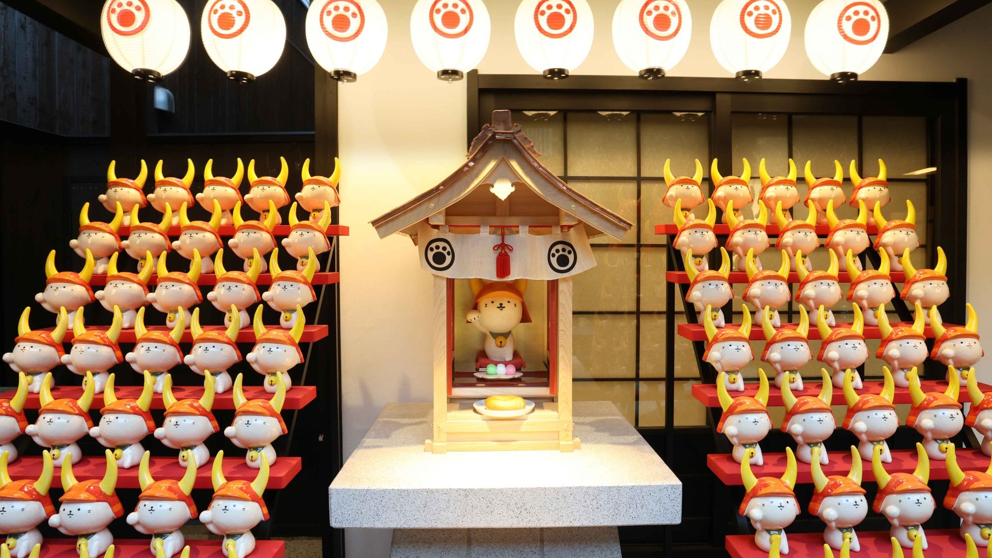 Una exhibición de figuritas dispuestas en filas, una estructura pequeña similar a un santuario con una figura en su interior, y faroles tradicionales arriba.