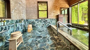 Una cabaña de madera con baño de baldosas de piedra, un taburete de madera y una ventana con vistas al follaje.
