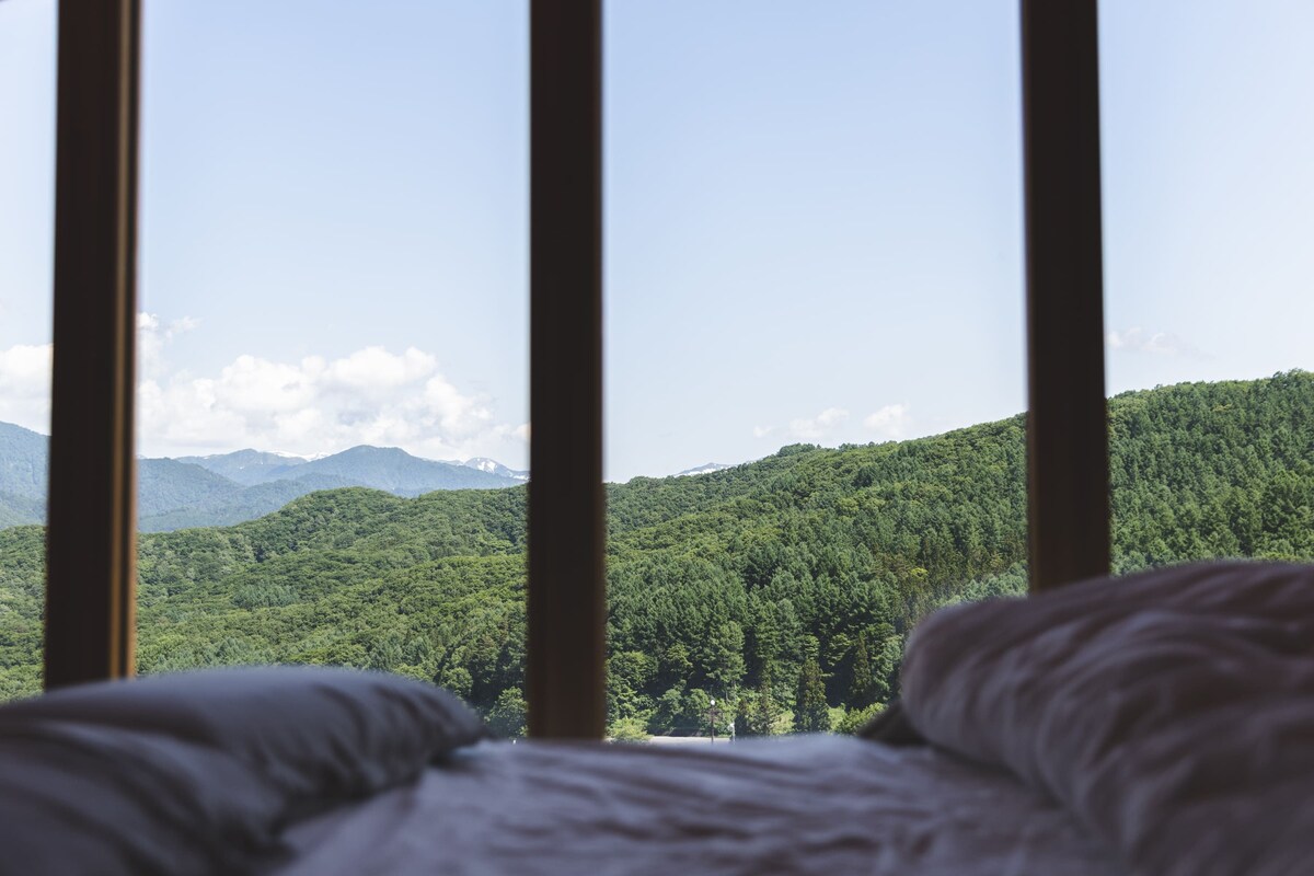 A bed with a view of a forested mountain range through a window.