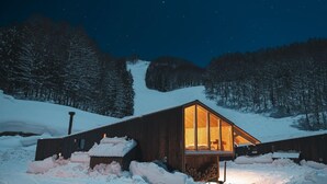 Eine Holzhütte mit einem großen Glasfenster, umgeben von verschneitem Gelände und Bäumen.