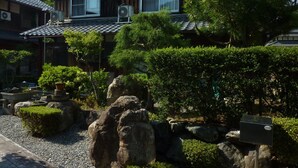 A traditional Japanese house with a tiled roof, surrounded by greenery and rocks.