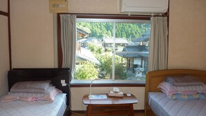 A room with two beds, a small table, an air conditioning unit, and a window with a view of traditional buildings.