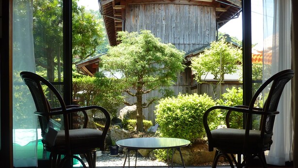 A room with two wicker chairs and a small table, a wooden deck, and a view of a traditional wooden structure and greenery outside.