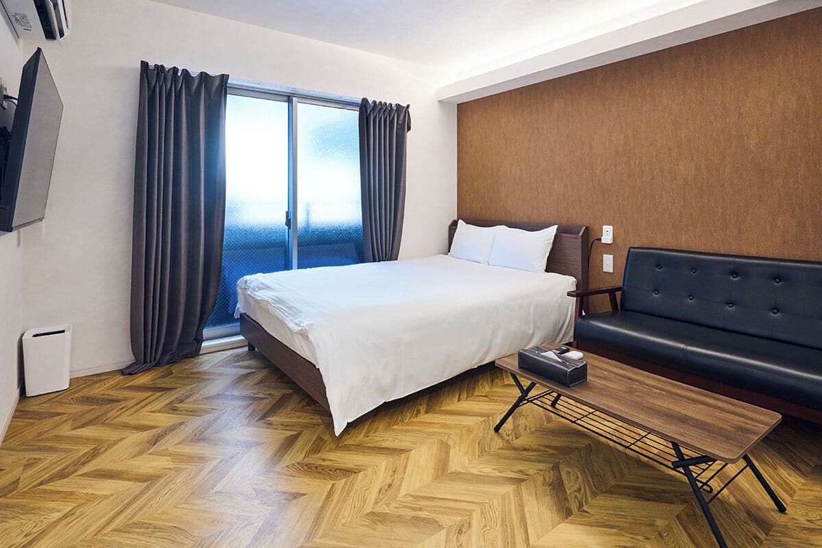 Quarto de hotel com cama, sofá, piso de madeira, janela com cortinas e televisão na parede.