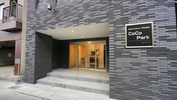 The entrance to an apartment hotel with a modern facade, a sign indicating it's a CoCo Park, and a set of stairs leading to the entrance.