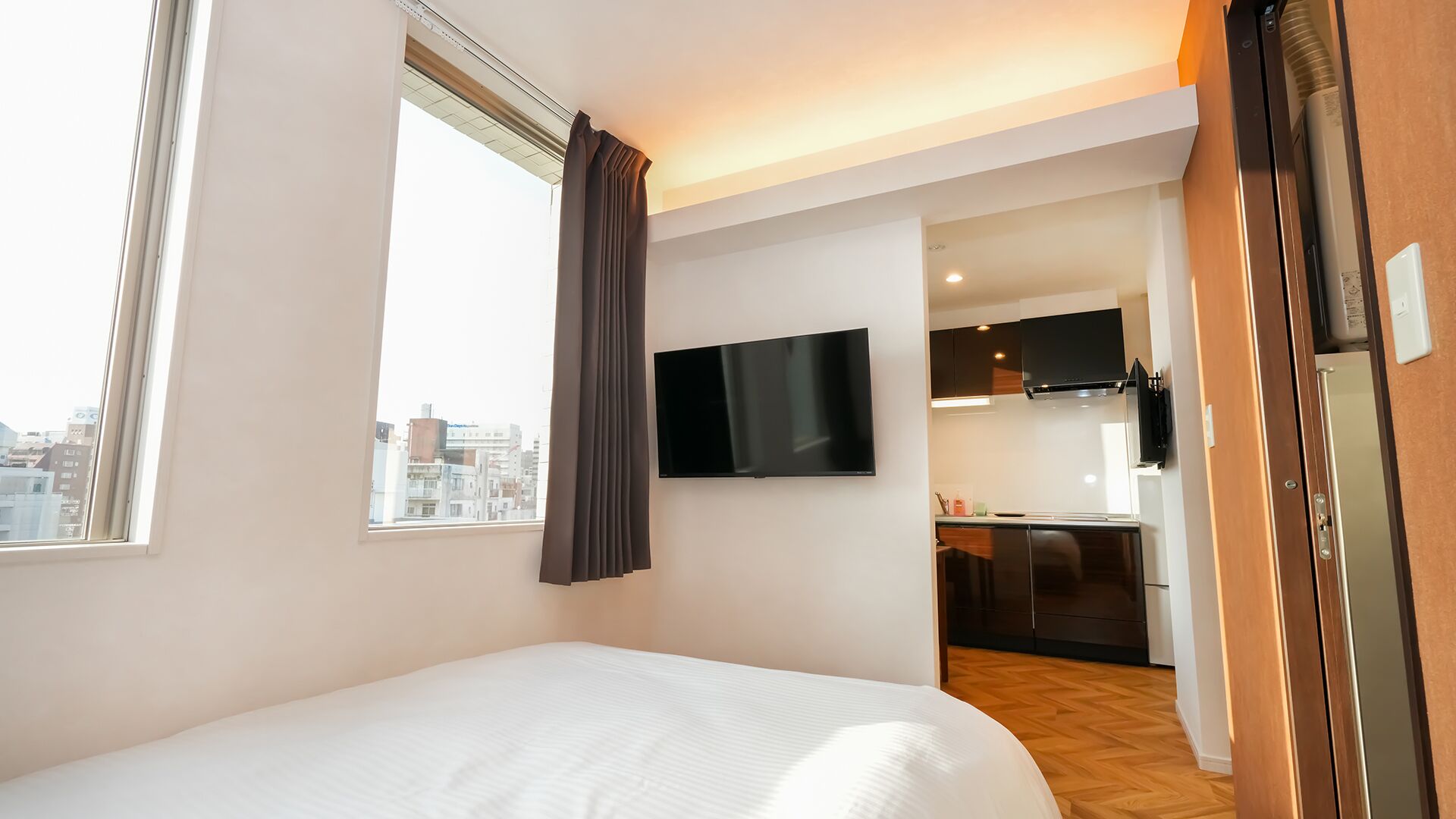 A hotel room with a bed, a TV mounted on the wall, a window with curtains, and a view of the cityscape.