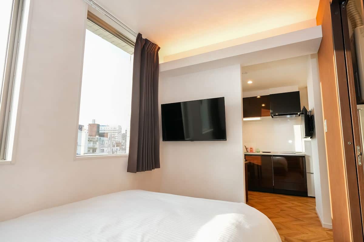 Quarto de hotel com cama, televisão presa à parede, janela com cortinas e vista para a paisagem urbana.