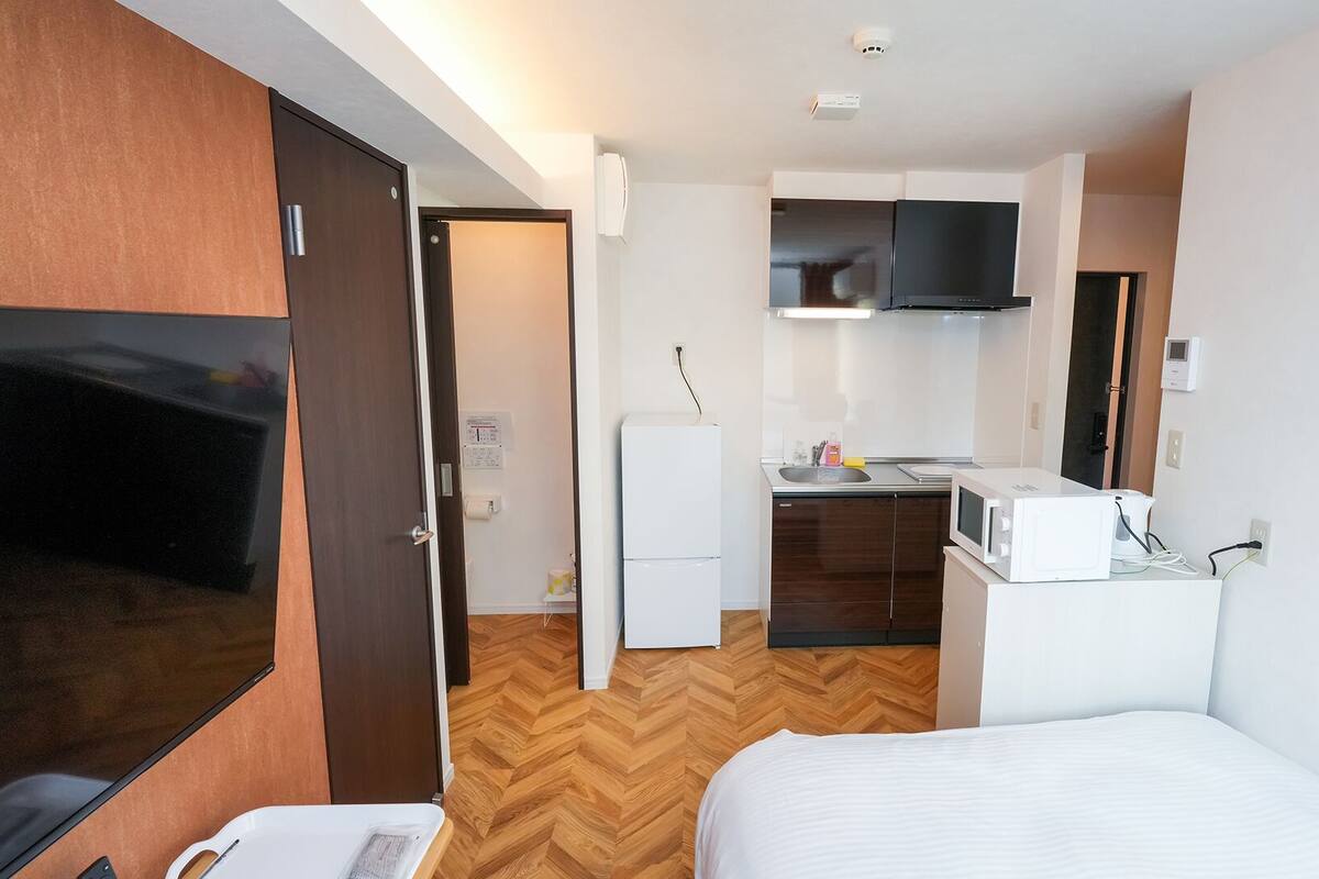 Quarto de hotel com cama, televisão, cozinha compacta e piso de madeira.