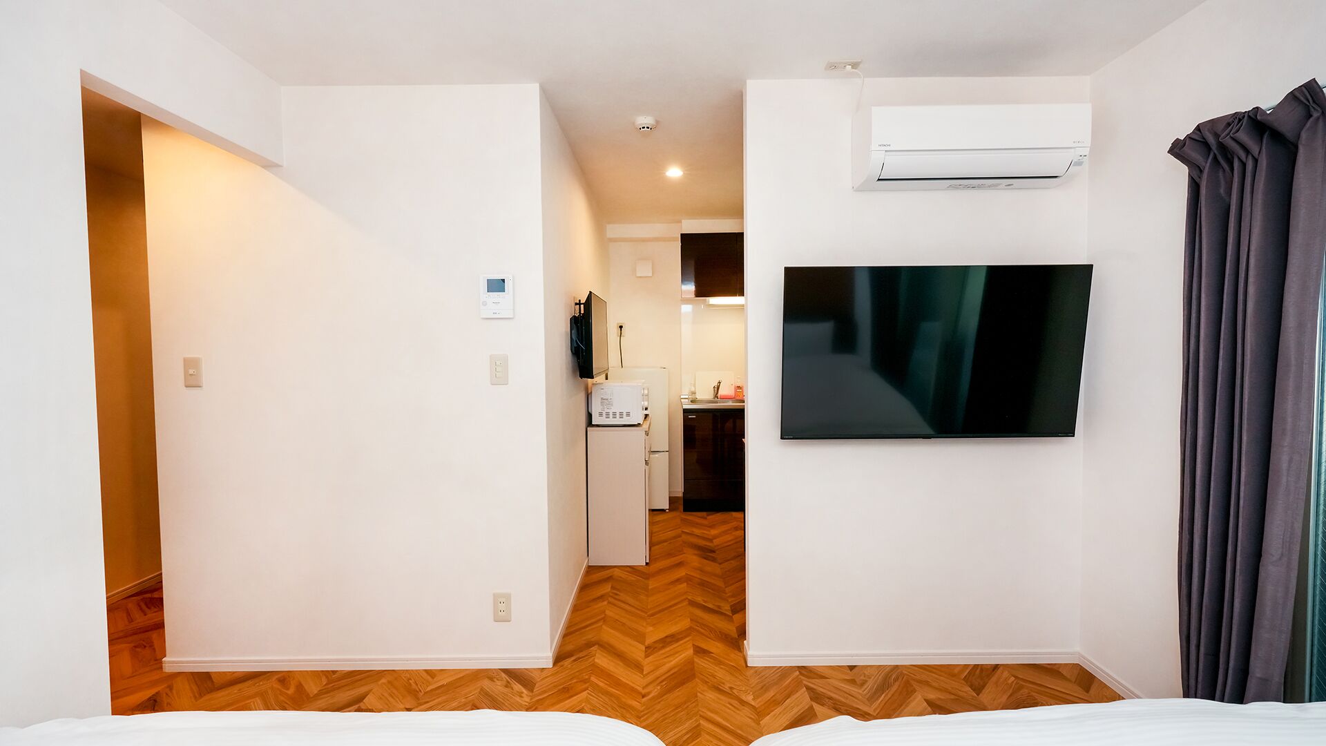 A hotel room with a bed, a TV, an air conditioner, and a kitchen area in the background.