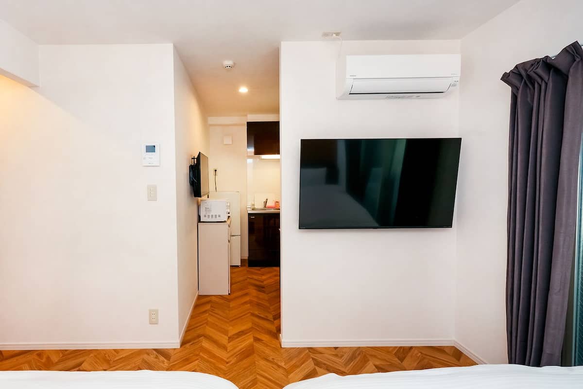 Quarto de hotel com cama, televisão, ar condicionado e área de cozinha ao fundo.