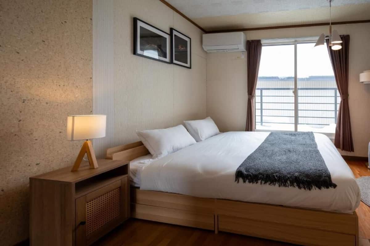 Ein ordentlich gemachtes Bett mit weißen Laken und einer dunkelgrauen Tagesdecke, ein Nachttisch mit Lampe sowie ein Fenster mit Vorhängen.