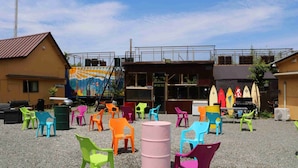 An outdoor seating area with colorful plastic chairs and tables, a large barrel, and surfboards in the background.