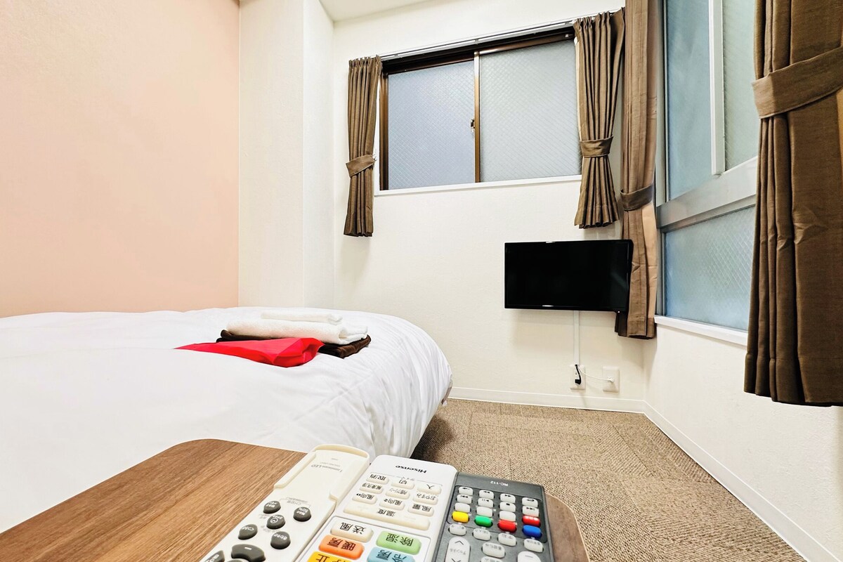 Una habitación de hotel con cama, televisor montado en la pared y control remoto sobre la mesita de noche.