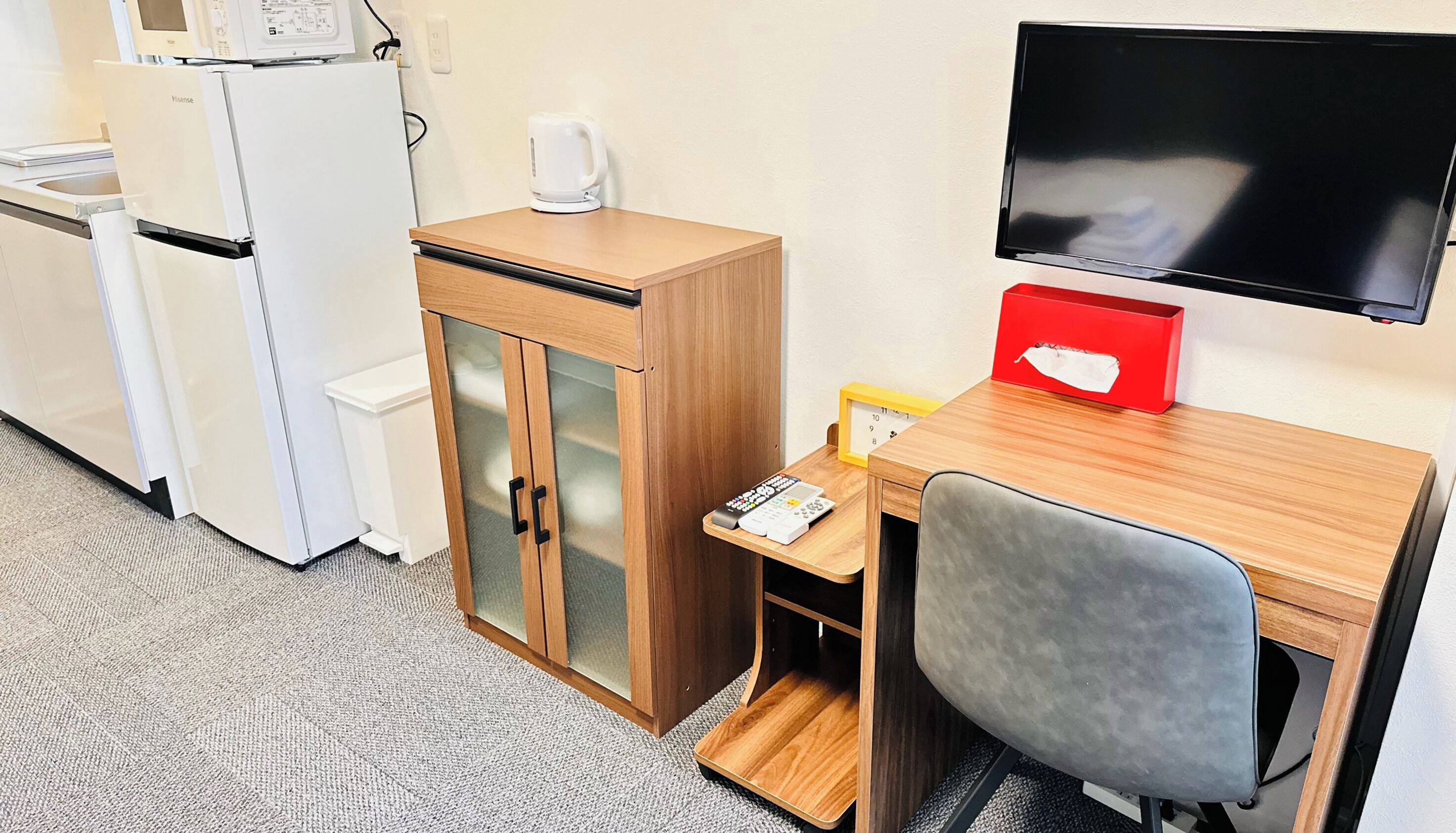A compact hotel room with a kitchenette, a wooden desk with a chair, a television, and a small red box.