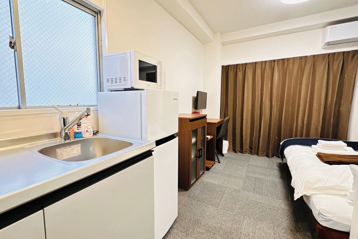 Habitación de hotel compacta con kitchenette, una cama individual, un escritorio y una ventana con cortinas.
