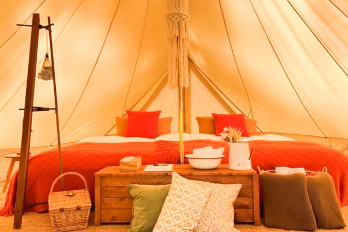 A tent with a bed, pillows, a table with a bowl, a basket, and a hanging light fixture.