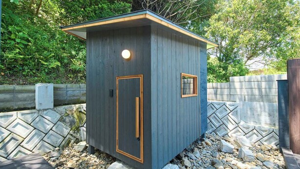 A small, dark-colored wooden shed with a door and a window, surrounded by a stone wall and greenery.