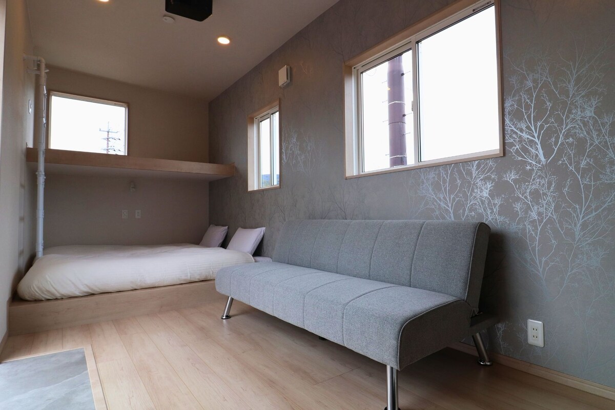 A modern room with a bunk bed, a grey sofa, large windows, and a patterned wall.