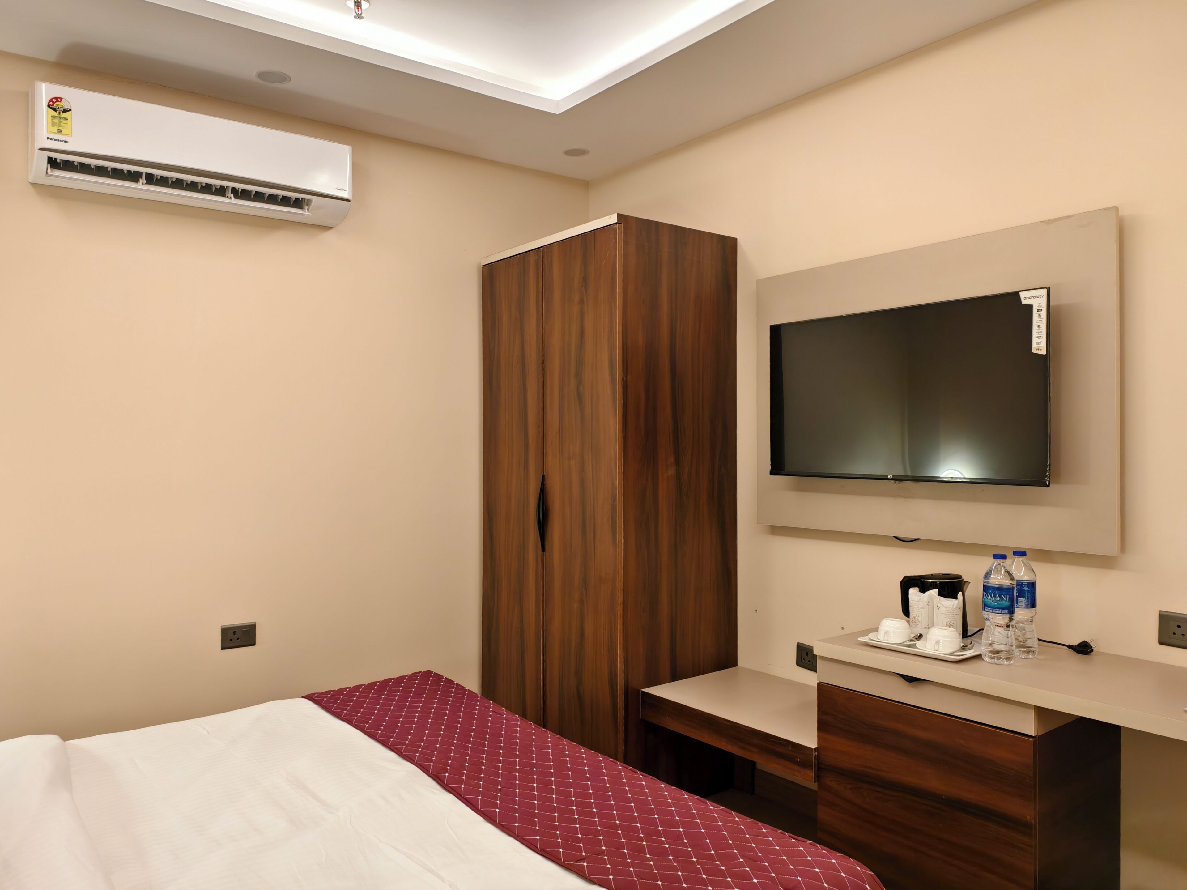Una habitación de hotel con una cama, un armario de madera, un televisor montado en la pared, un escritorio con una taza y una botella, y un aire acondicionado.
