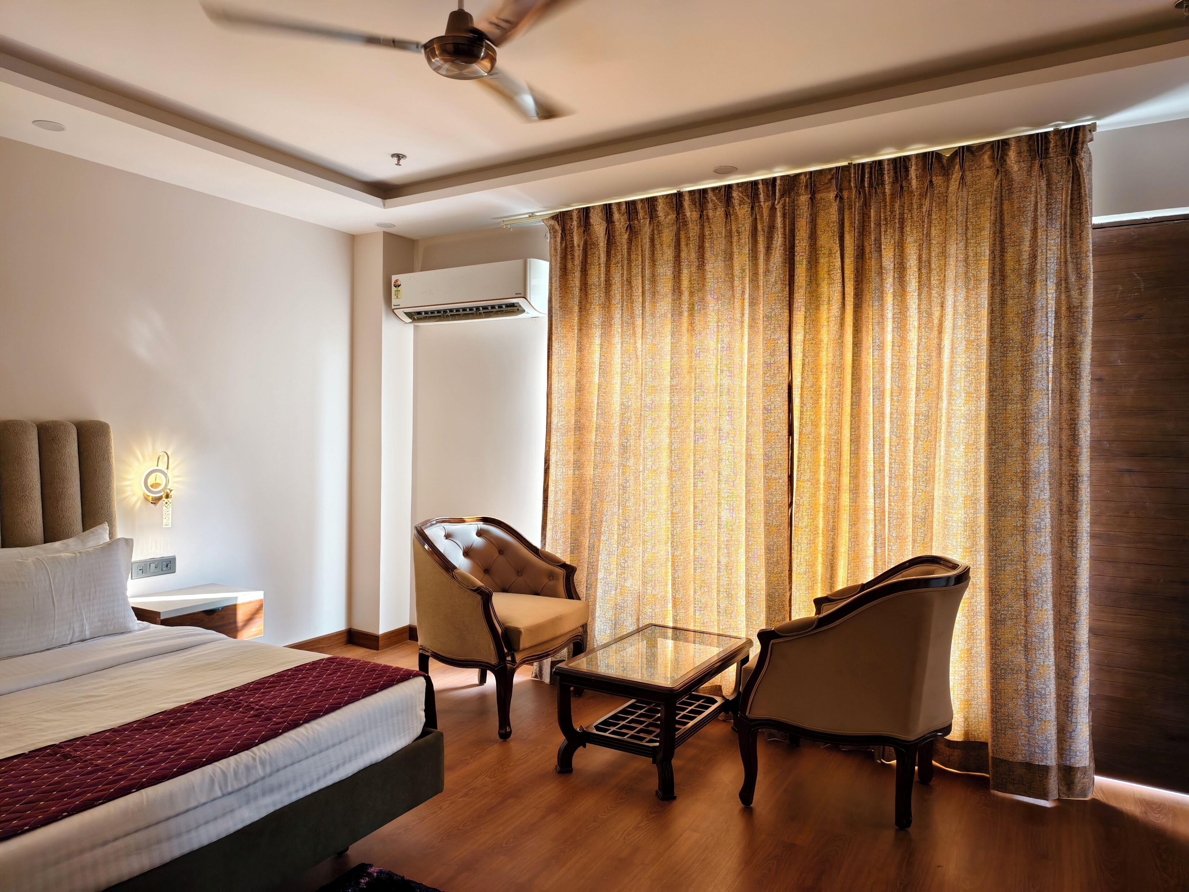 Habitación de hotel con cama, dos sillas, una mesita, ventilador de techo y ventana con cortinas.