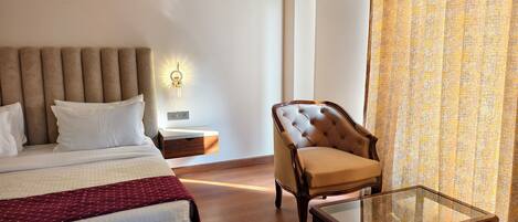 Habitación de hotel con cama, sillón, mesita pequeña, unidad de aire acondicionado y mesita de noche.