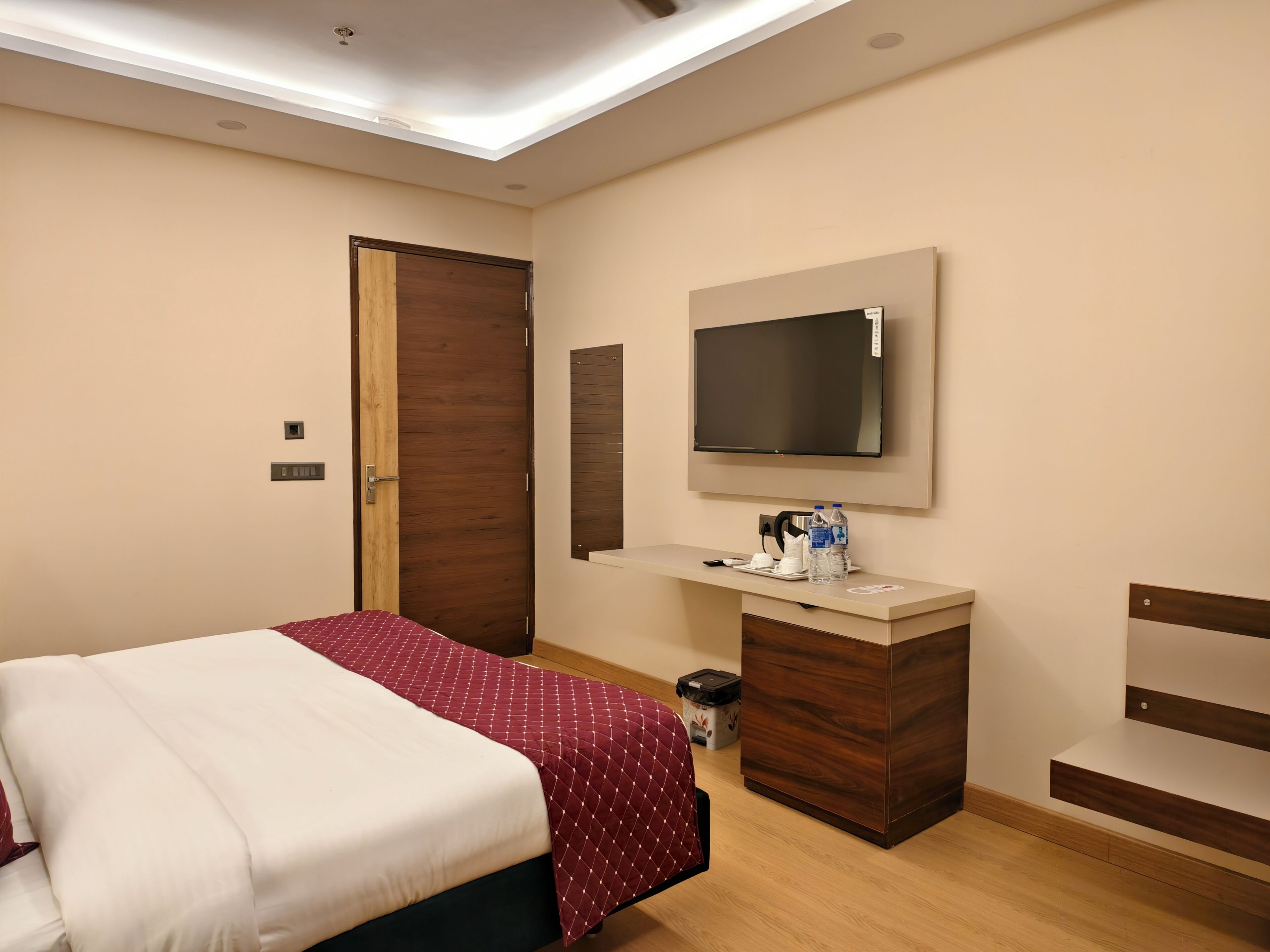 Een slaapkamer met een bed, een aan de muur bevestigde televisie, een bureau met spiegel en een houten deur.