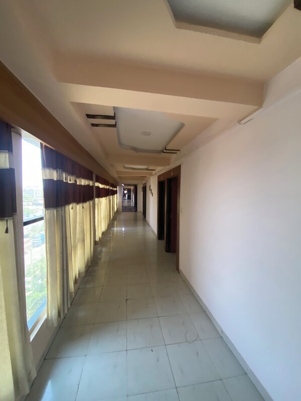 A long, narrow hallway with a tiled floor, a ceiling with recessed lighting, and windows with curtains on one side. - Hotel Anand Palace Inn (Ahmedabad)