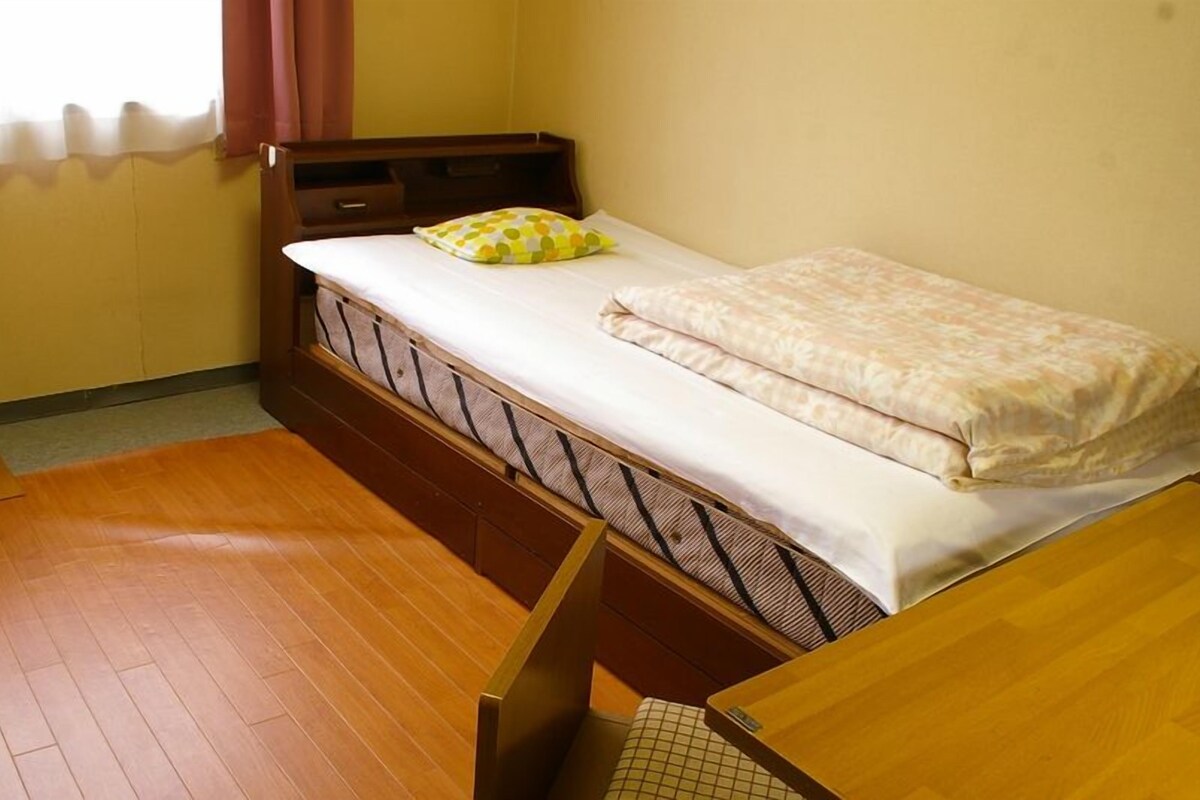 A single bed with a mattress, a pillow, a wooden headboard, a small bedside table, and a television on a desk.