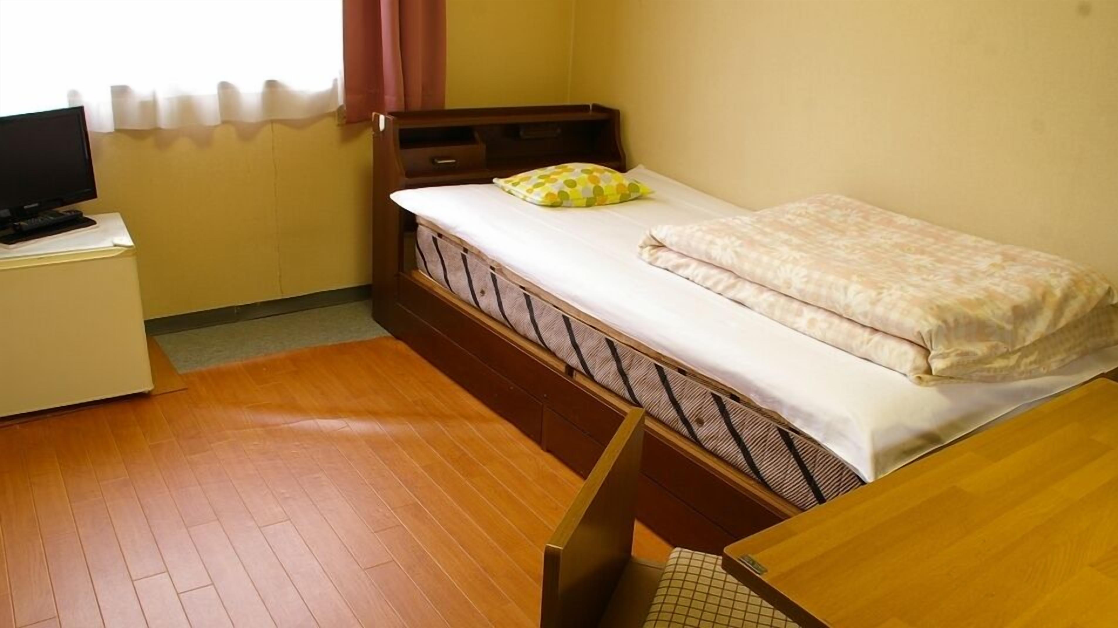 A single bed with a mattress, a pillow, a wooden headboard, a small bedside table, and a television on a desk.