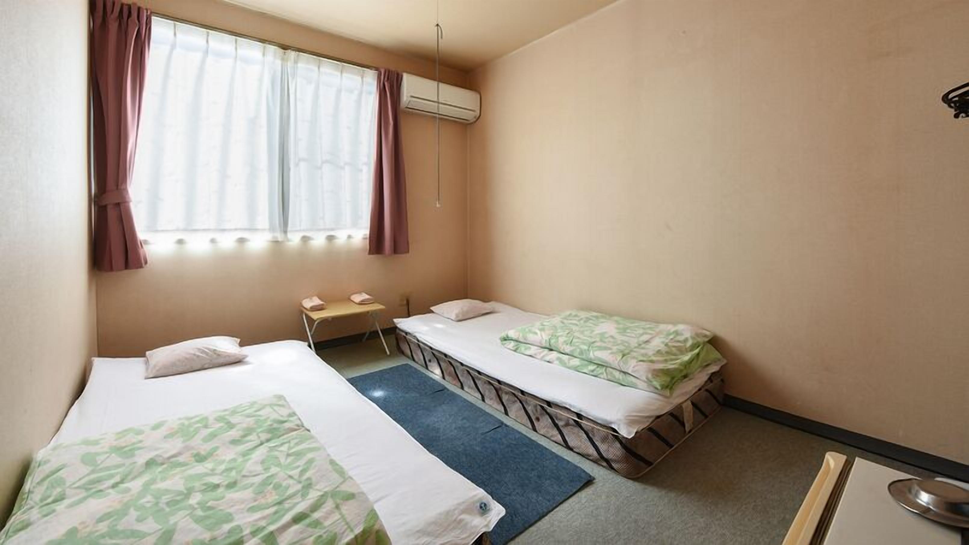 A small, single-room hotel room with two beds, a window with curtains, an air conditioning unit, and a small table.