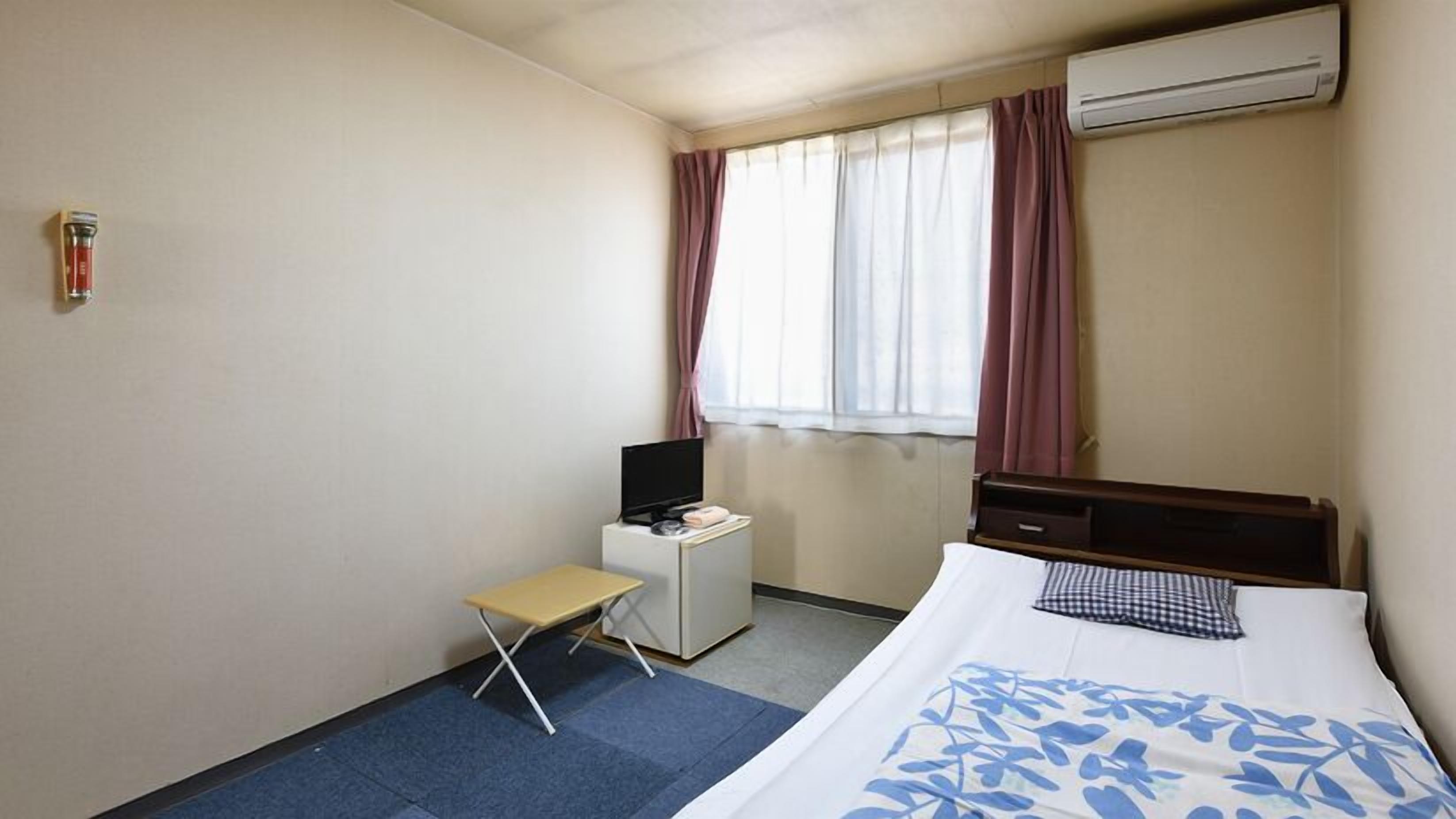 A hotel room with a bed, a desk with a TV, a small table, and an air conditioning unit.
