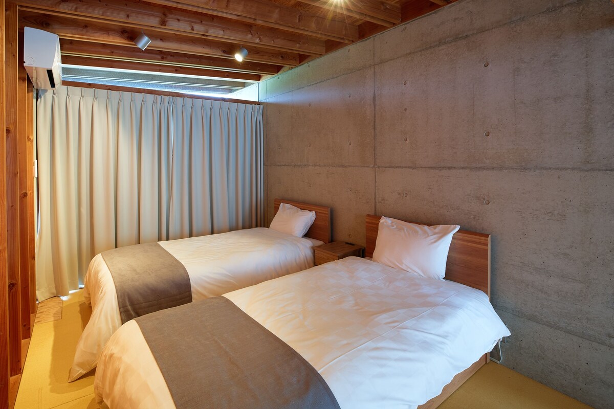 Una habitación con dos camas, vigas de madera en el techo, una pared de hormigón y una ventana con cortinas.