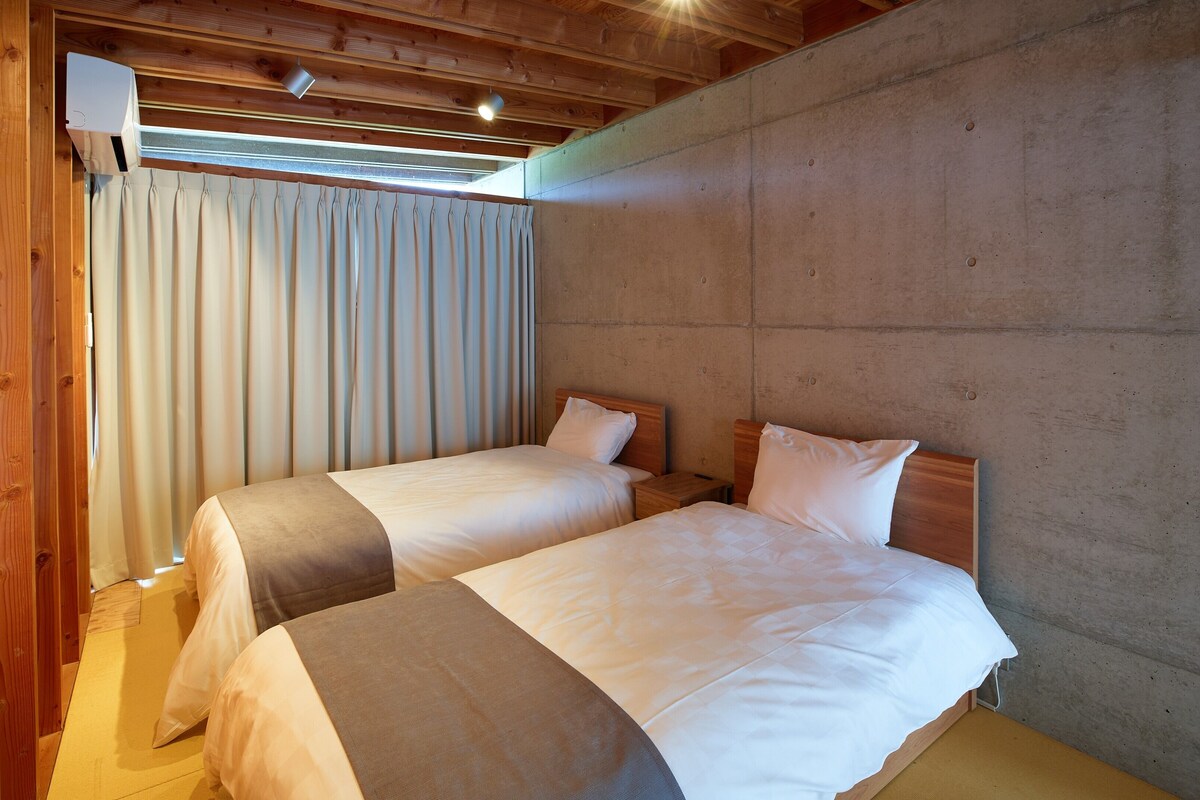 Een kamer met twee bedden, houten balkenplafond, een betonwand en een raam met gordijnen.