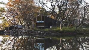 Una cabaña junto al lago con pared de piedra, terraza de madera y reflejo en el agua.