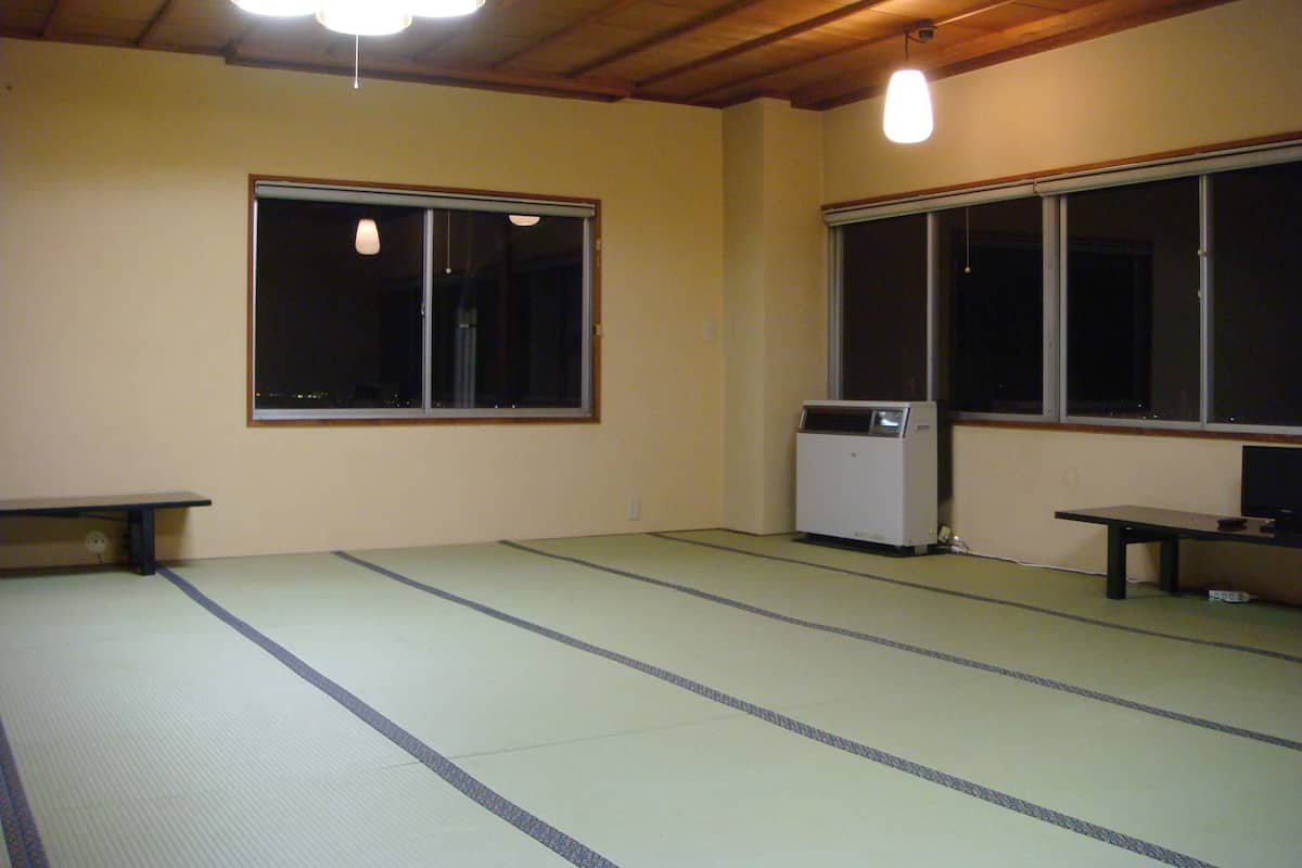 A room with tatami flooring, a heater, a bench, and two windows.