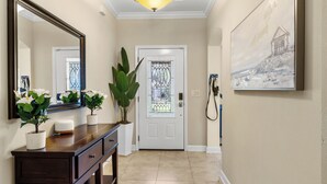 Un pasillo con una consola de madera, plantas en macetas, un espejo grande, una puerta con vidriera y un cuadro en la pared.