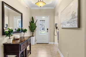 Un couloir avec une console en bois, des plantes en pot, un grand miroir, une porte ornée d’un vitrail et un tableau accroché au mur.