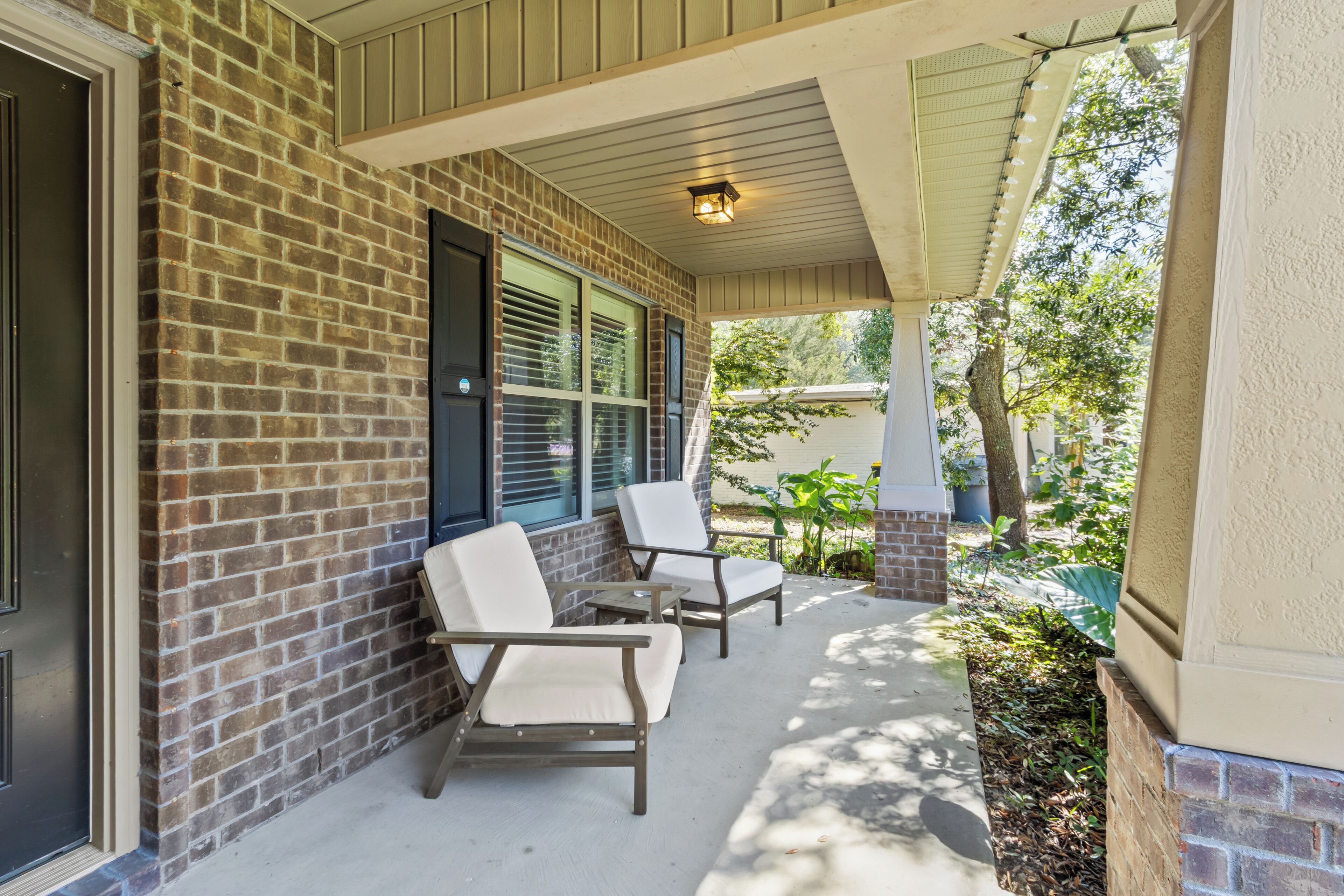 A covered porch with brick walls, two white cushioned chairs, and a small table.