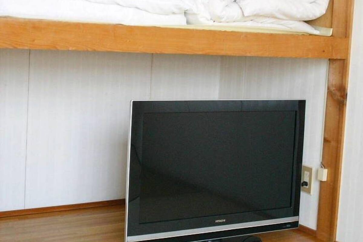 Una habitación con televisión sobre un soporte, un estante de madera con mantas dobladas y una pared blanca.